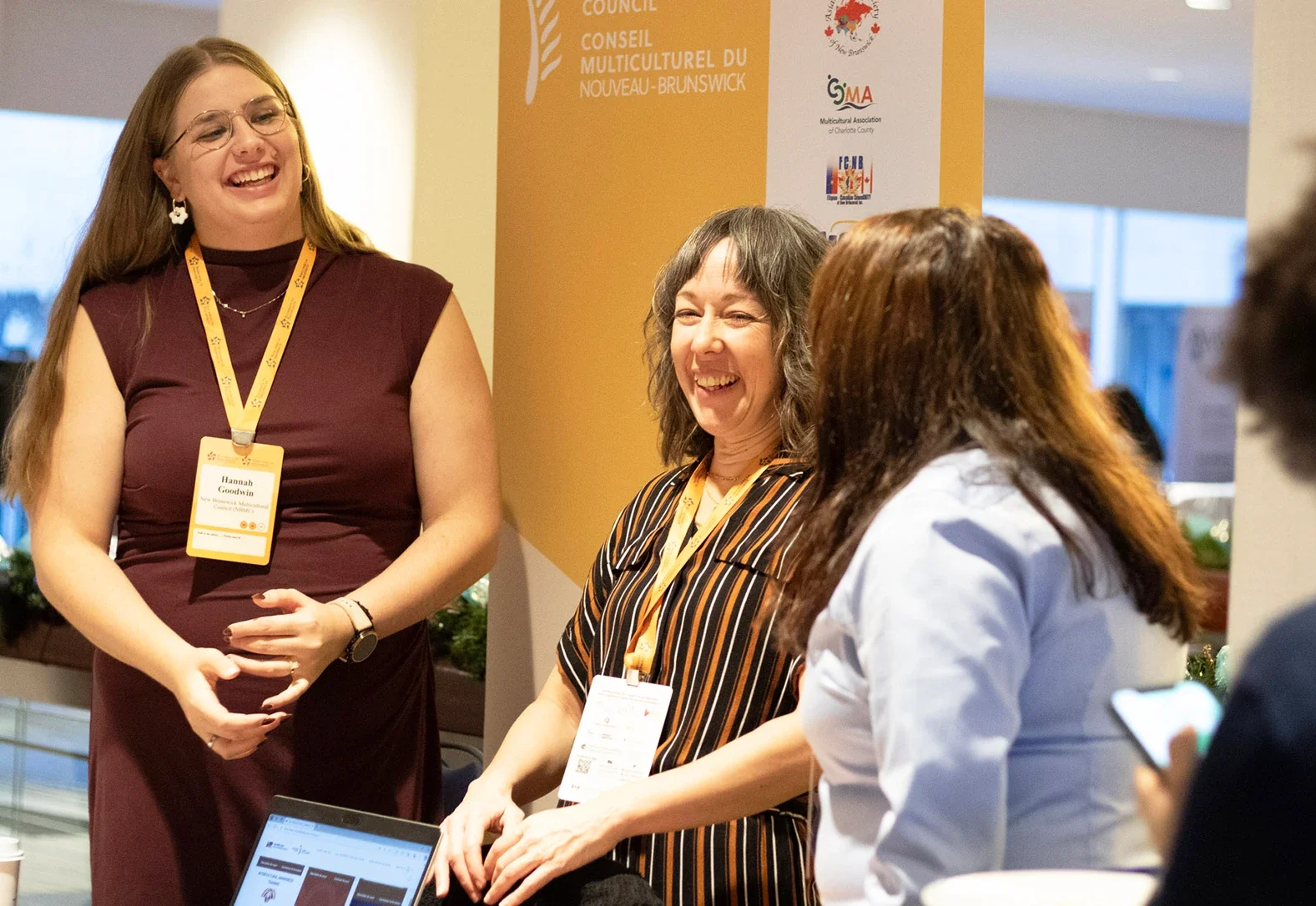 Three conference attendees laughing together in front of a New Brunswick Multicultural Council banner.
