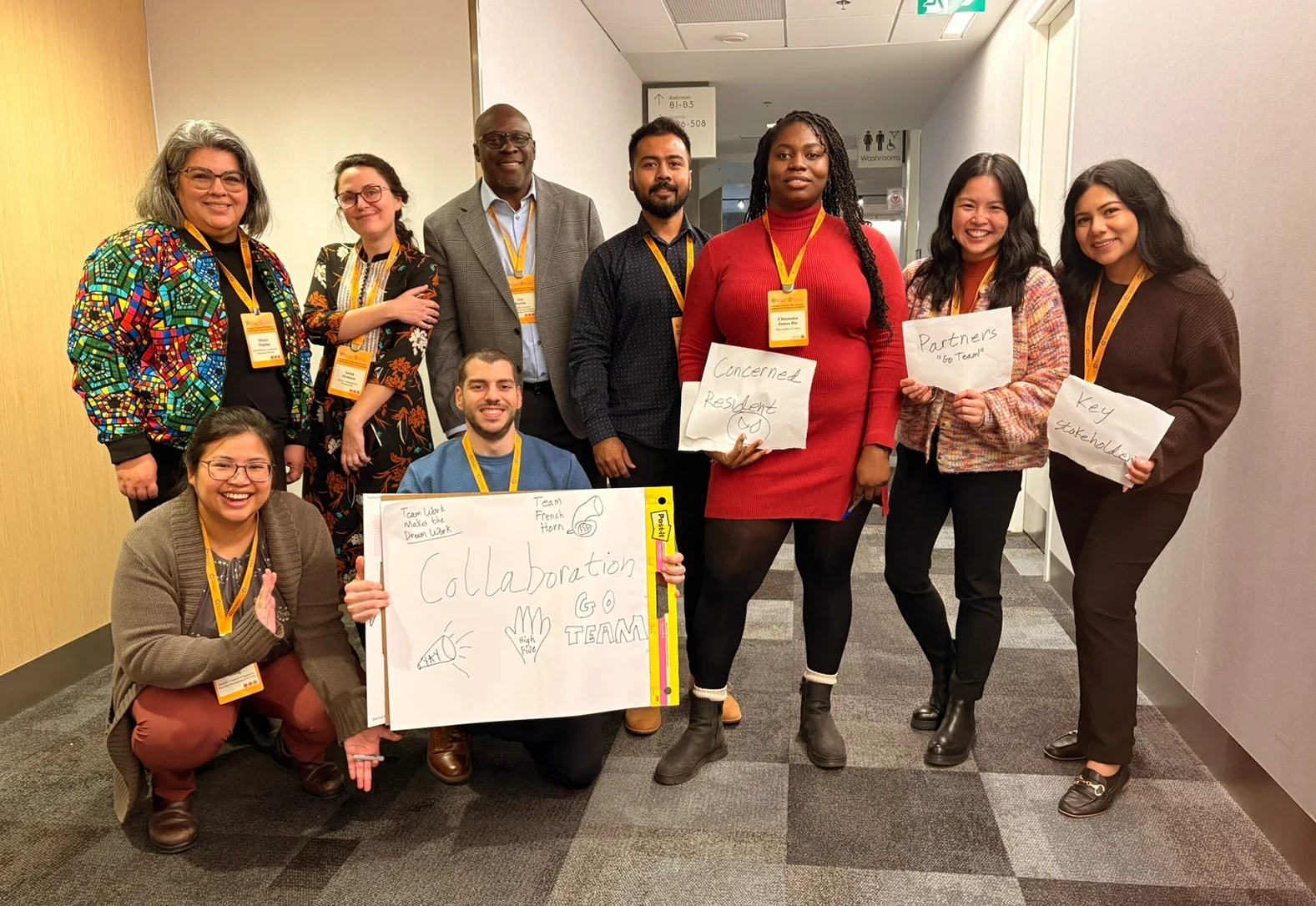 Nine conference participants posing in a hallway holding handwritten signs reading "Collaboration Go Team", "Concerned Resident", "Partners" and "Key Stakeholder".
