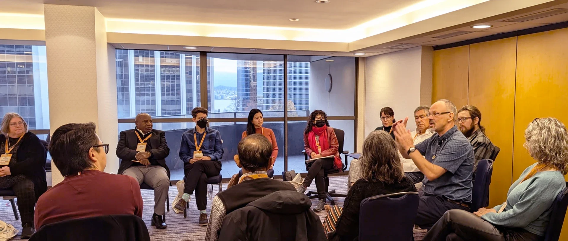 Conference participants seated in a circle in a meeting room, one speaking with the group listening.